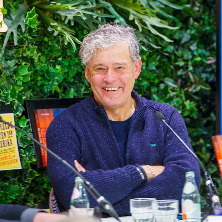 Charles Groenhuijsen lachend aan een debattafel met microfoons, met zijn boeken over Amerika op de achtergrond