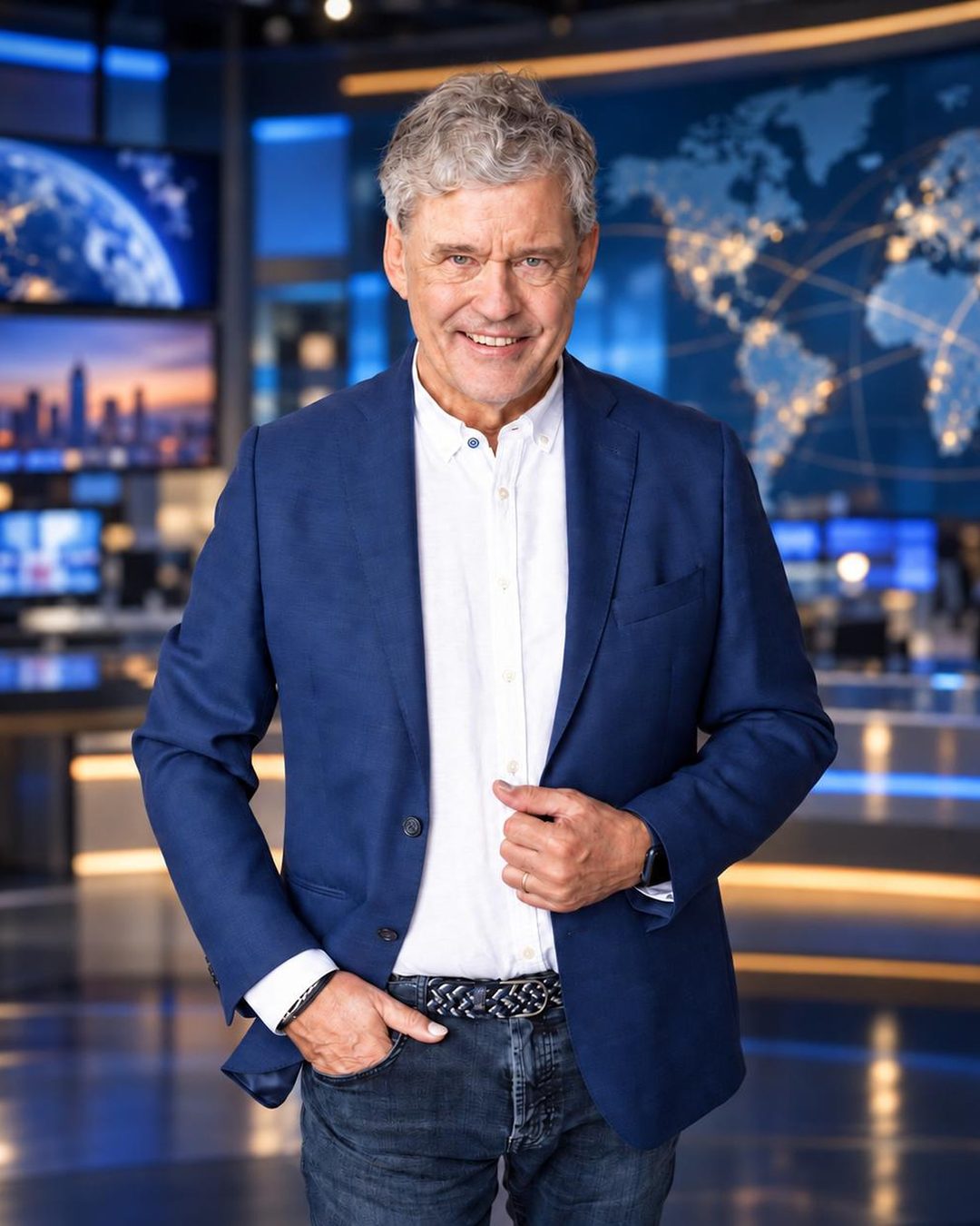 Charles Groenhuijsen in blauw colbert in een televisiestudio, met nieuwsschermen en een verlichte wereldkaart achter hem